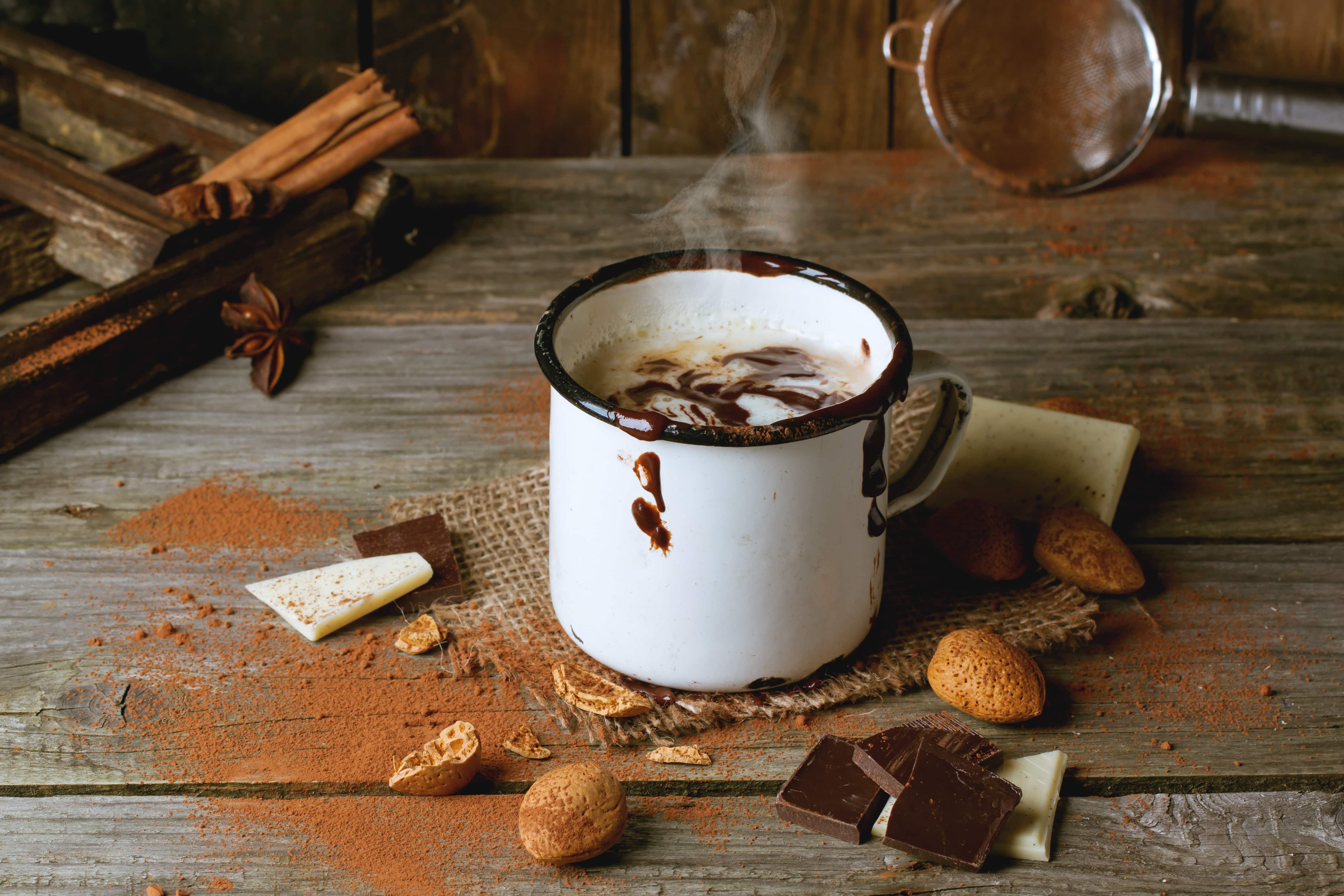 Vintage mug with hot chocolate served with chunks of white and dark chocolate and almonds on old wooden table