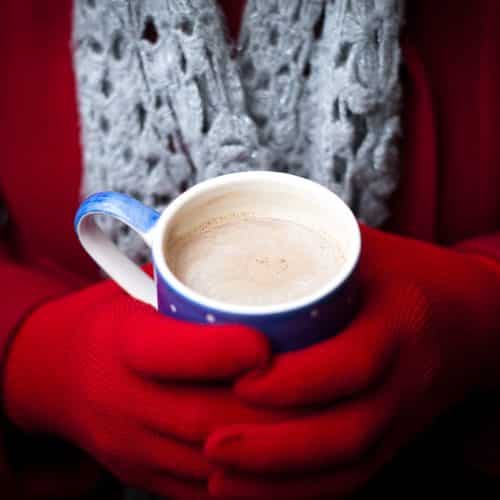 A woman in a red coat and gloves warms her hands on a mug of hot chocolate.  Focus on the drink.