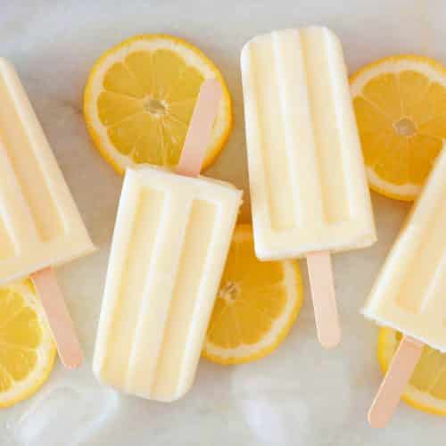 Healthy lemon yogurt popsicles with fresh lemon slices scattered on a white marble background