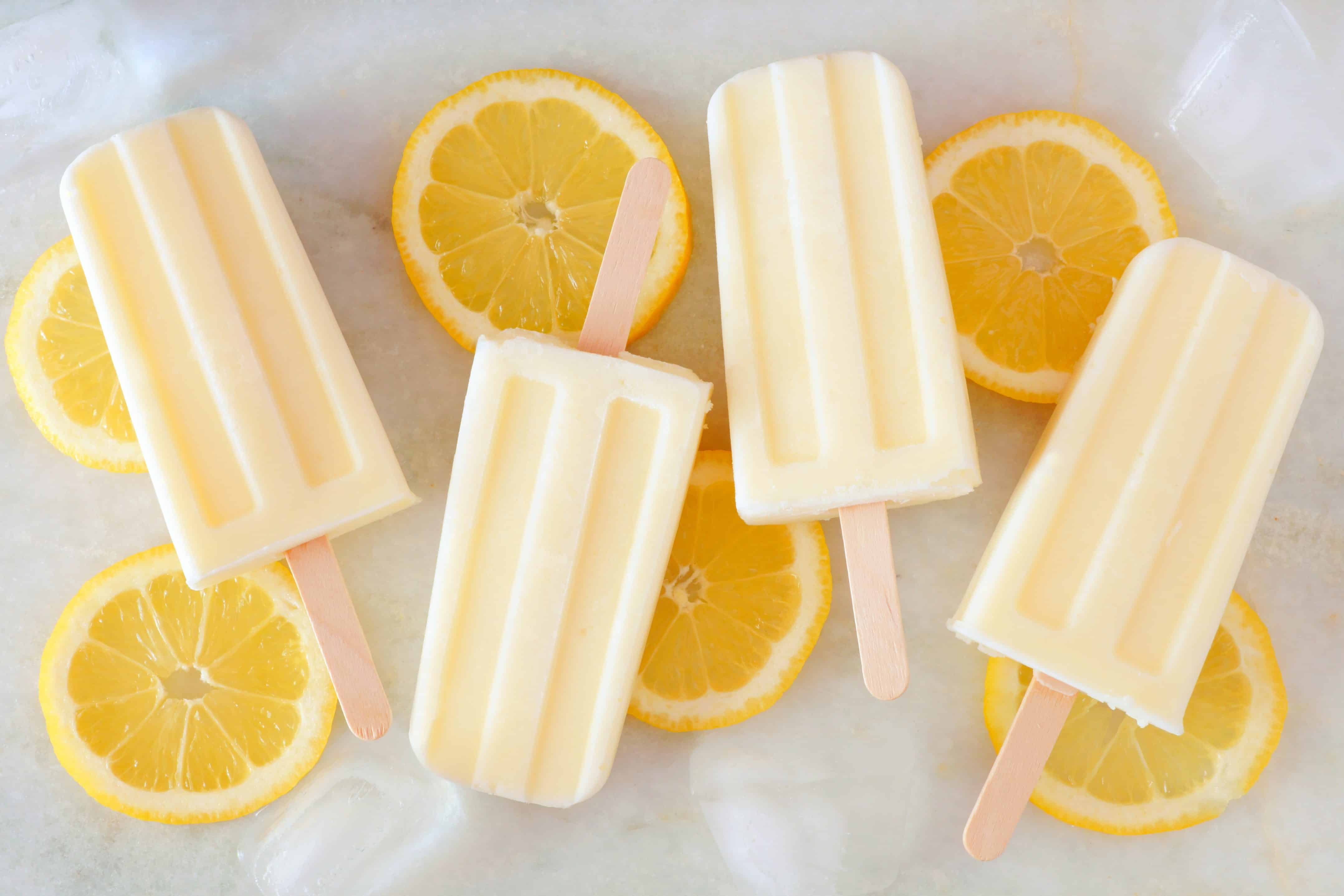 Healthy lemon yogurt popsicles with fresh lemon slices scattered on a white marble background